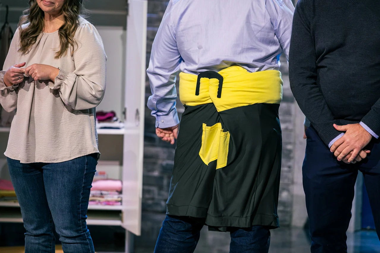 Drei Personen stehen vor einem offenen Schrank. Die mittlere Person trägt eine gelbe Schärpe und eine schwarze Schürze.