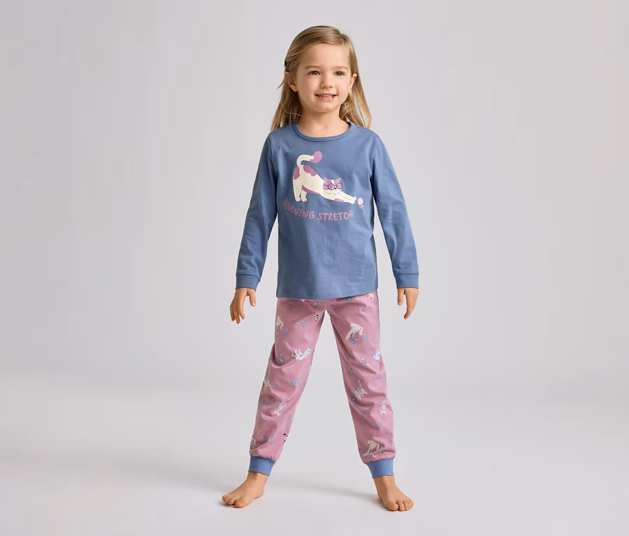 Ein junges Mädchen steht barfuß und lächelt in einem blauen Langarmshirt mit einer streckenden Katze und rosa Hosen mit Einhornmuster.