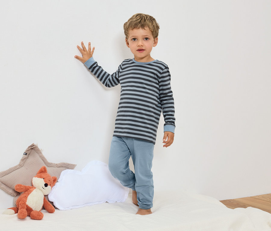Ein Junge steht in blau gestreiftem Pyjama neben einem Fuchs Stofftier und Kissen.
