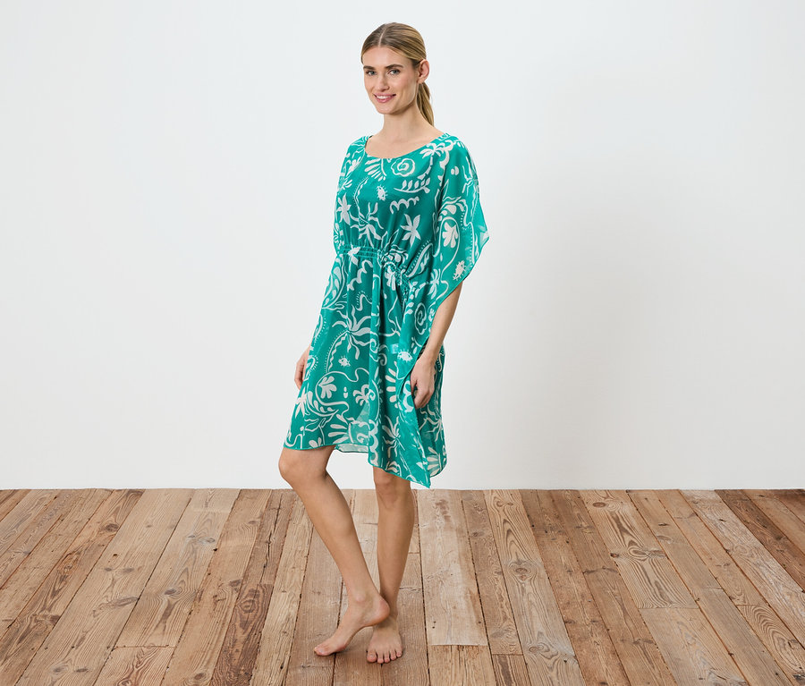 Frau steht barfuß auf Holzboden, trägt ein grünes Kleid mit weißem Blumenmuster und einem Kordelzug in der Taille.
