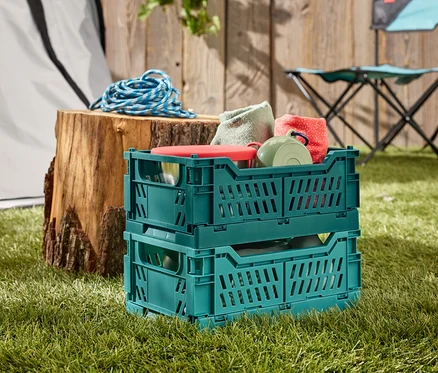 Zwei grüne Klappboxen stehen gestapelt auf Gras, gefüllt mit Geschirr und Textilien. Im Hintergrund ein Baumstumpf mit blauem Seil und ein Campingstuhl.