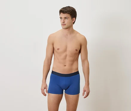 Mann mit freiem Oberkörper und blauen Boxershorts vor einem weißen Hintergrund.