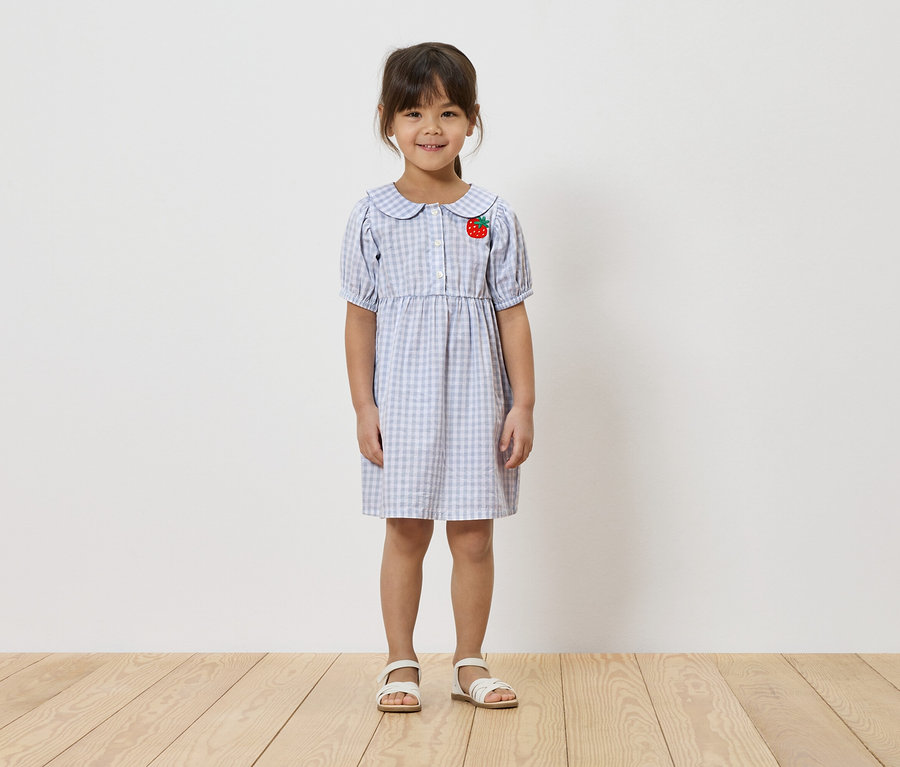 Mädchen trägt ein hellblau kariertes Kleid mit Erdbeer-Applikation und weiße Sandalen.