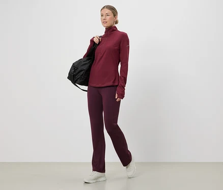 Frau in weinrotem Sportoberteil und Hose mit schwarzer Tasche über der Schulter, weiße Turnschuhe.