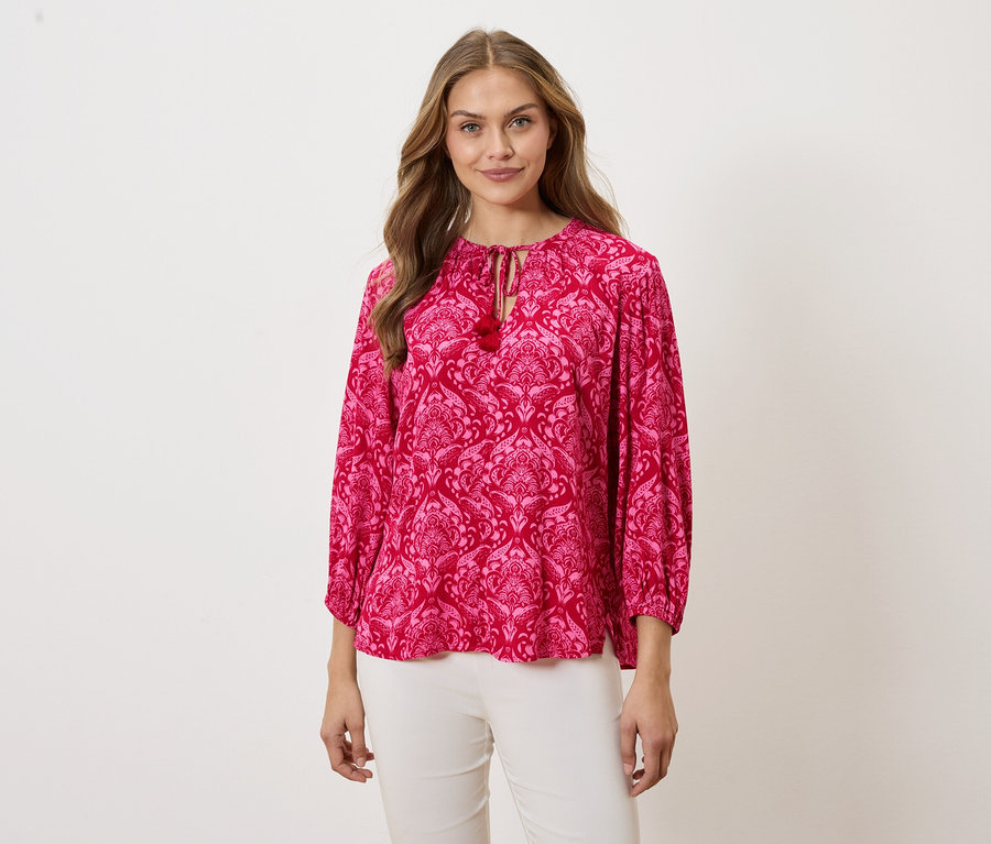 Frau trägt eine rosafarbene Bluse mit Muster und weiße Hosen.