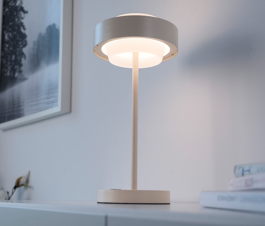Moderne Tischlampe mit rundem, geschichtetem Schirm, die ein warmes Licht abgibt, auf einer weißen Oberfläche neben einem gerahmten Schwarz-Weiß-Landschaftsbild.