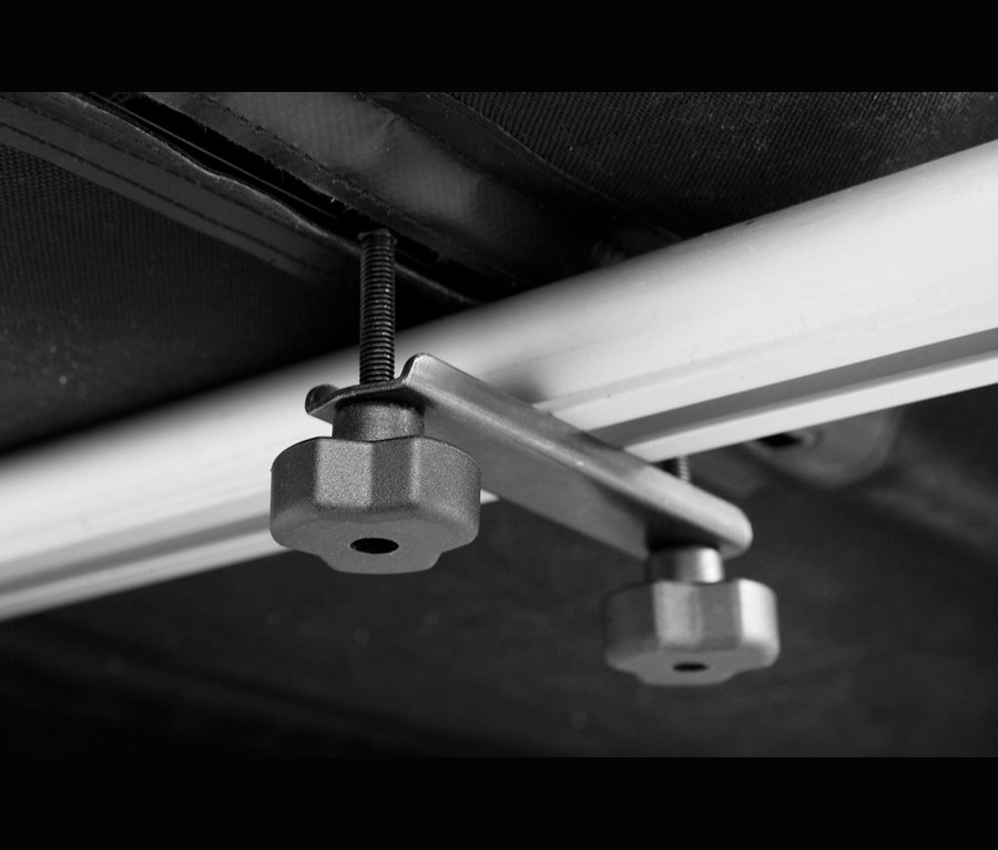 Nahaufnahme eines schwarz-weißen Klemmmechanismus mit einer Gewindeschraube, einer Metallhalterung und zwei sechseckigen Knöpfen, der ein dunkles Material an einer hellen Schiene befestigt.