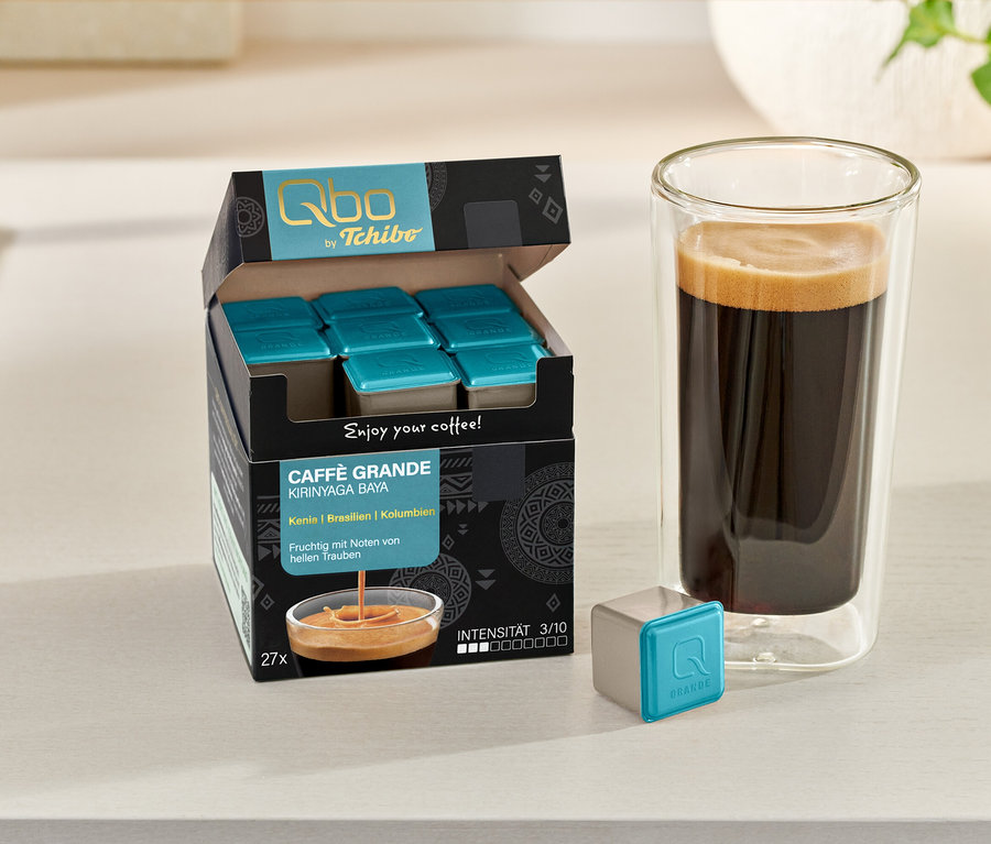 Eine geöffnete Packung Qbo Caffè Grande KIRINYAGA BAYA Kapseln, ein Glas Kaffee und eine einzelne Kapsel sind auf einem Tisch.