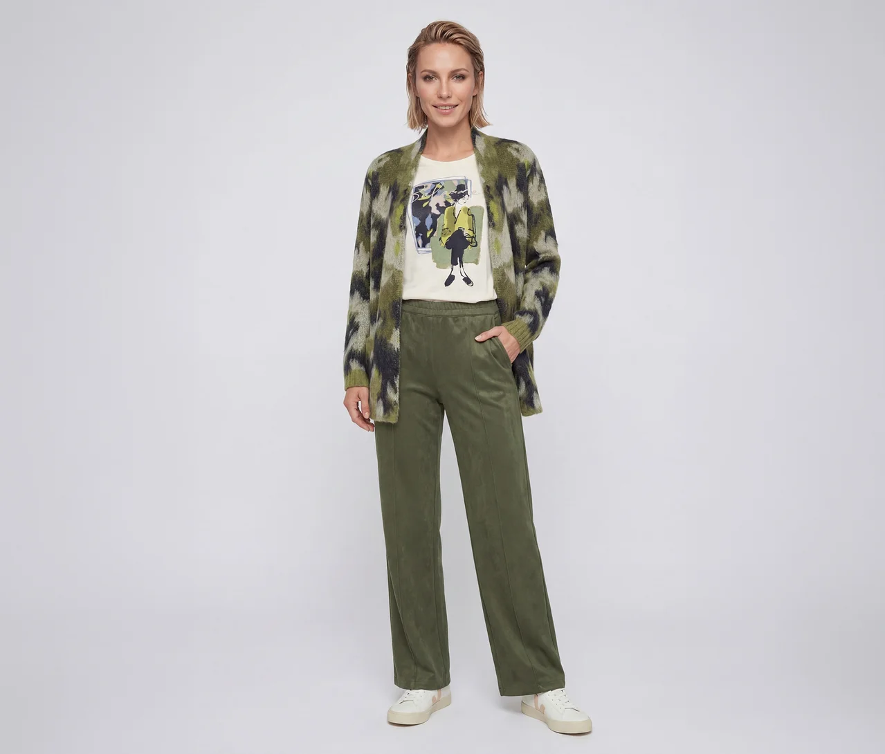 Lächelnde Frau trägt eine grün gemusterte Strickjacke, ein helles T-Shirt mit Grafikdruck, olivgrüne Hose und weiße Sneaker.