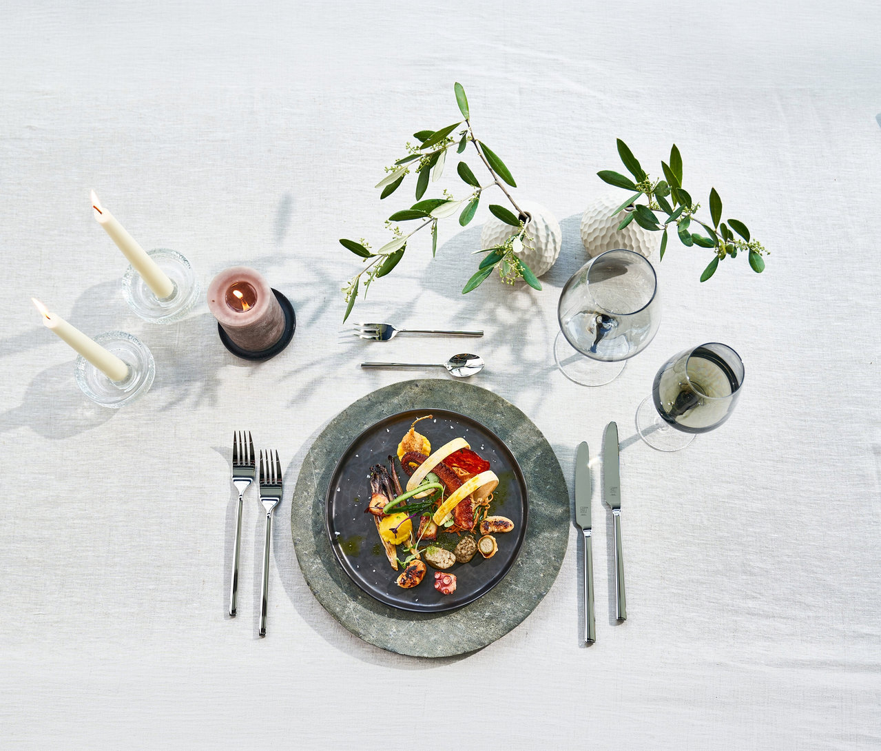 Ein gedeckter Tisch mit einem ZWILLING "Aberdeen" Besteck-Set, das Gabeln, Messer und einen Löffel umfasst. Ein Teller mit Essen, Kerzen, Weingläser und Olivenzweige sind darauf arrangiert.