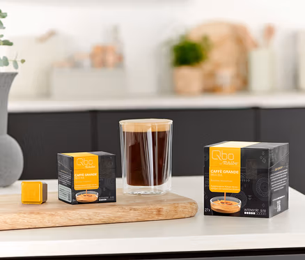 Ein Glas Kaffee steht neben einer Qbo Caffè Grande BELO SUL - 8 Kapsel Packung und einer einzelnen Kapsel auf einem Holzbrett.
