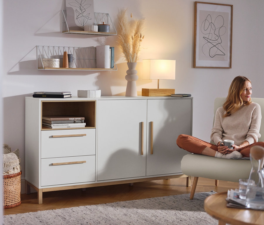 Frau sitzt im Wohnzimmer neben einem weißen Sideboard »Eklund« mit Schubladen und Tür.