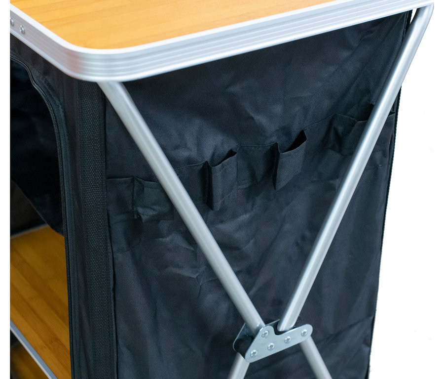 Nahaufnahme eines faltbaren Campingtisches mit Stauraum. Er hat eine hellbraune Platte, silberne Beine und schwarze Stoffseiten mit Taschen und Reißverschluss.