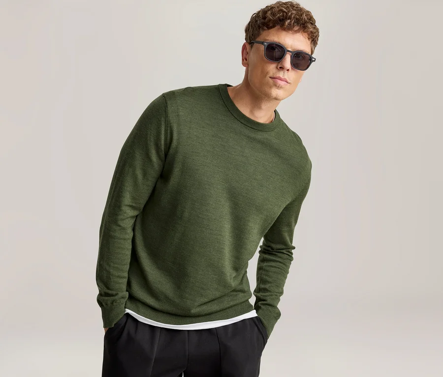 Mann mit lockigem braunem Haar trägt ein grünes Langarmshirt, schwarze Hose und dunkle Sonnenbrille, steht mit Händen in den Hosentaschen und blickt in die Kamera.