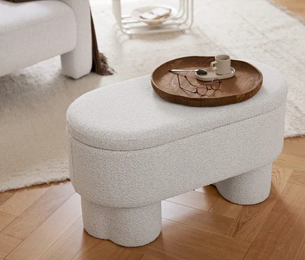 Ein hellgrauer, strukturierter Hocker mit einem Holztablett, auf dem eine Tasse, ein Löffel und eine Brille liegen. Daneben steht ein Sessel mit einer braunen Decke auf einem hellen Teppich und Holzboden.