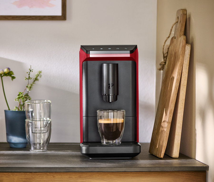 Roter und schwarzer Kaffeevollautomat mit einer Tasse Kaffee auf einer Arbeitsplatte, daneben Gläser und Schneidebretter.