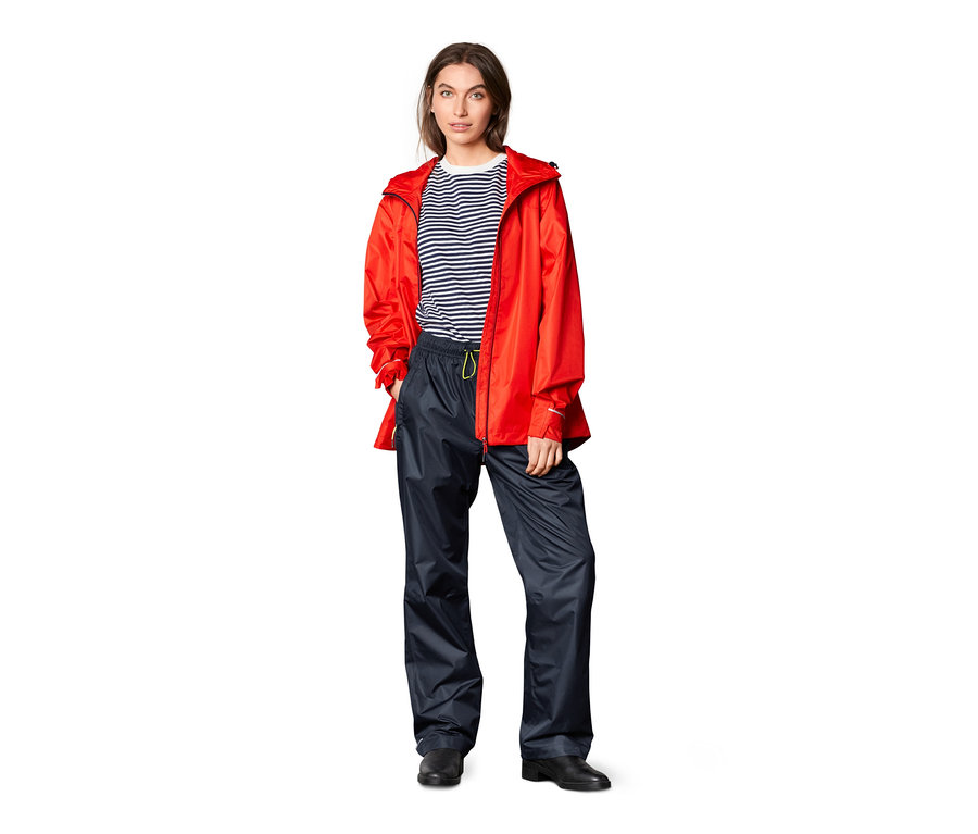 Eine Frau steht vor einem weißen Hintergrund und trägt eine Unisex-Regenhose und eine rote Jacke.