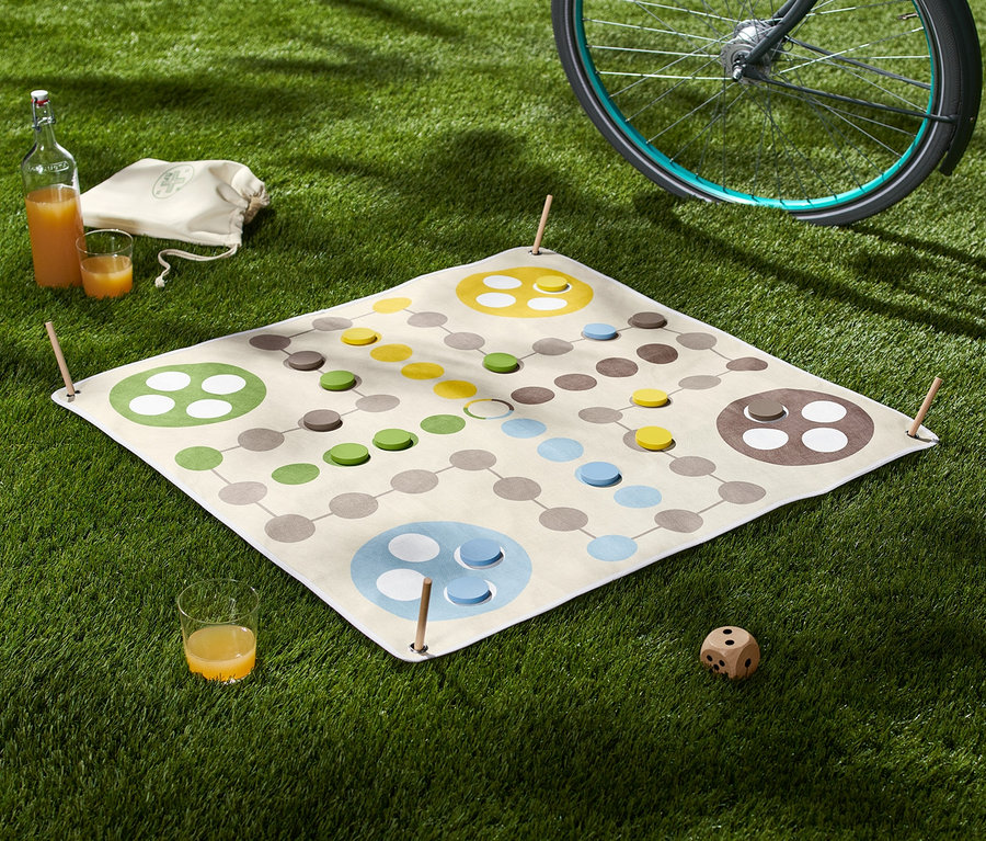 Auf dem Rasen liegt ein XL-Ludo-Spiel mit Würfel, daneben ein Glas Saft und eine Flasche. Im Hintergrund ein Fahrrad.