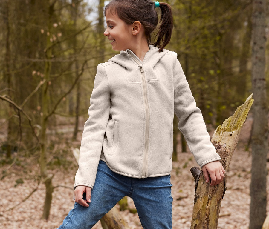 Ein Junge sitzt auf einem gefallenen Baum in gelben recycelbaren Gummistiefeln und einer braunen Wendemütze, dazu trägt er Joggdenim und eine Strickfleece-Jacke. Ein Mädchen steht auf dem Baum, trägt Joggdenim und eine Strickfleece-Jacke.