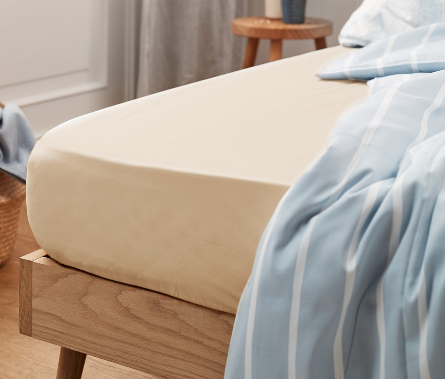 Beiges Jersey-Spannbettlaken Normalgröße auf einem Bett mit blau-weiß gestreifter Bettwäsche.