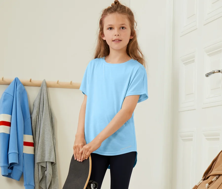 Mädchen hält ein Skateboard und trägt ein blaues Funktionsshirt und schwarze Leggings. Im Hintergrund sind 3 Mädchen-Funktionsshirts mit recyceltem Material zu sehen.