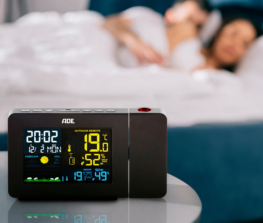 Auf einem Nachttisch steht eine ADE Wetterstation mit Funk-Außensensor und Projektion, im Hintergrund schläft ein Paar im Bett.