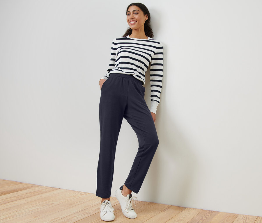 Frau lehnt an Wand, trägt marineblaue Joggpants und Streifenoberteil.