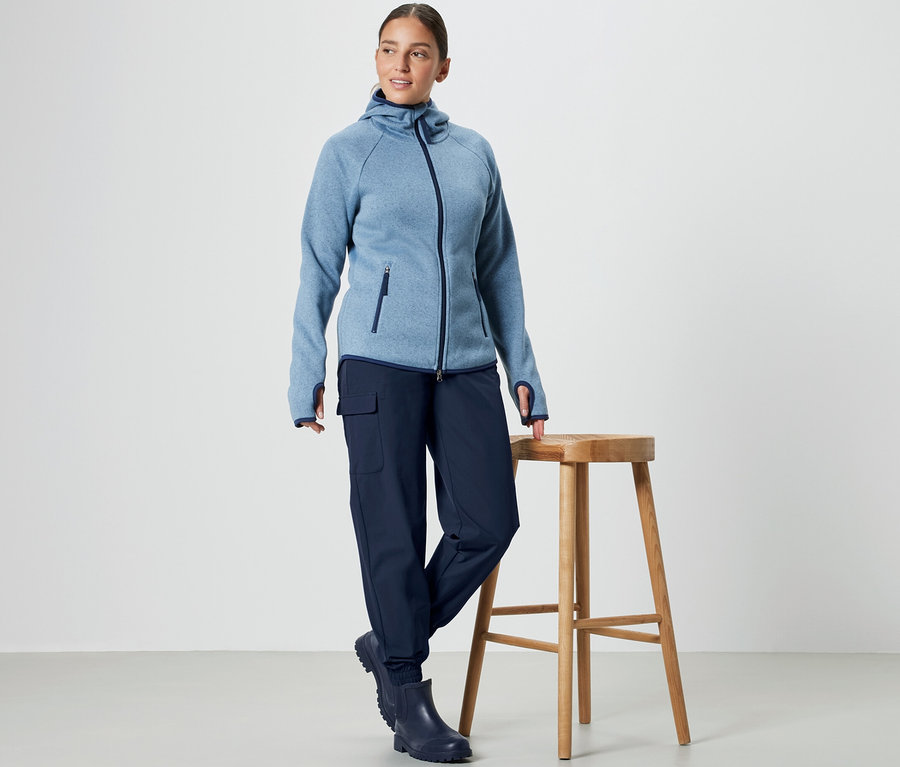 Eine Frau steht neben einem Holzhocker und trägt eine blaue Kapuzenjacke und eine Softshell-Joggpants.