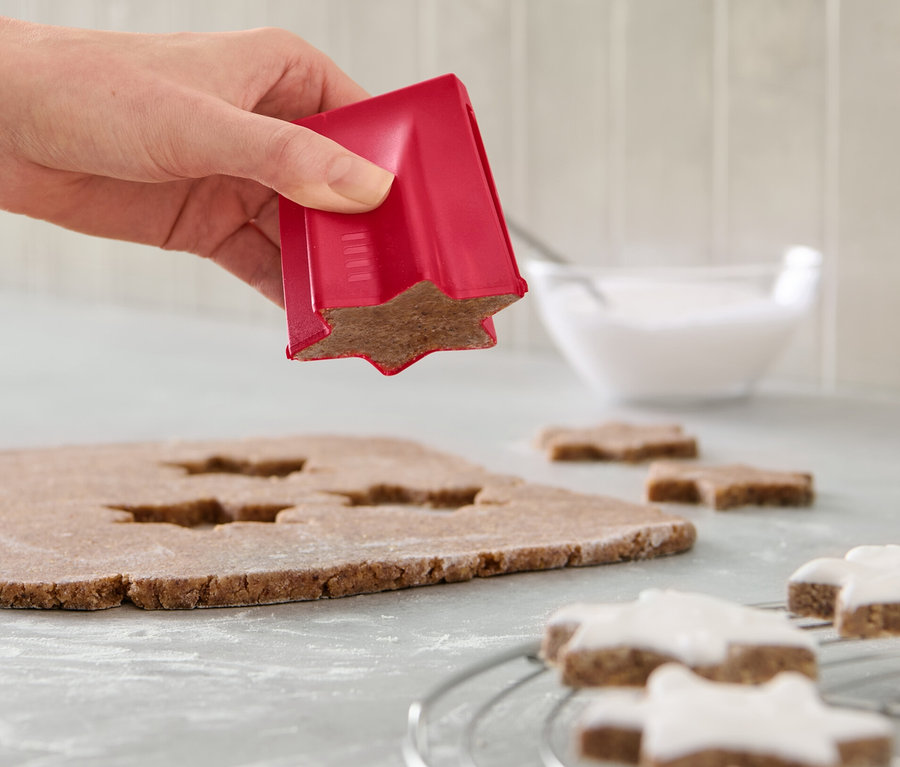 Eine Hand benutzt einen Zimtsternausstecher auf Teig. Auf der Theke liegen verzierte Sterne und eine Schüssel mit Zuckerguss.