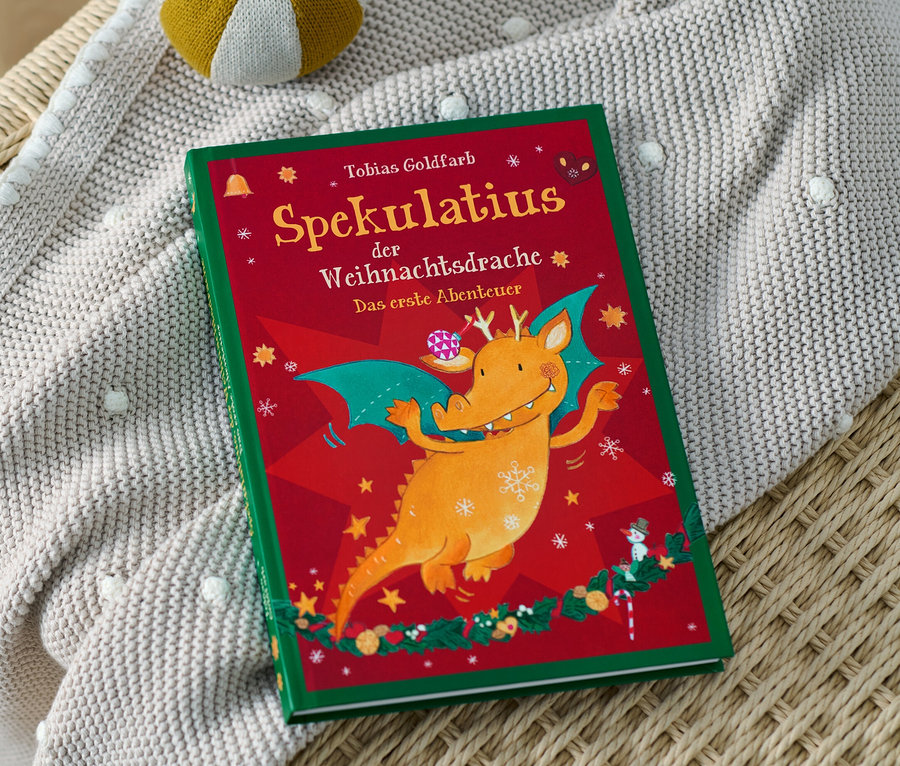 Auf einem Rattansessel liegen das Buch »Spekulatius, der Weihnachtsdrache« und eine Strickdecke mit Bommeln. Im Hintergrund ist ein gelb-weißes Spielzeug.