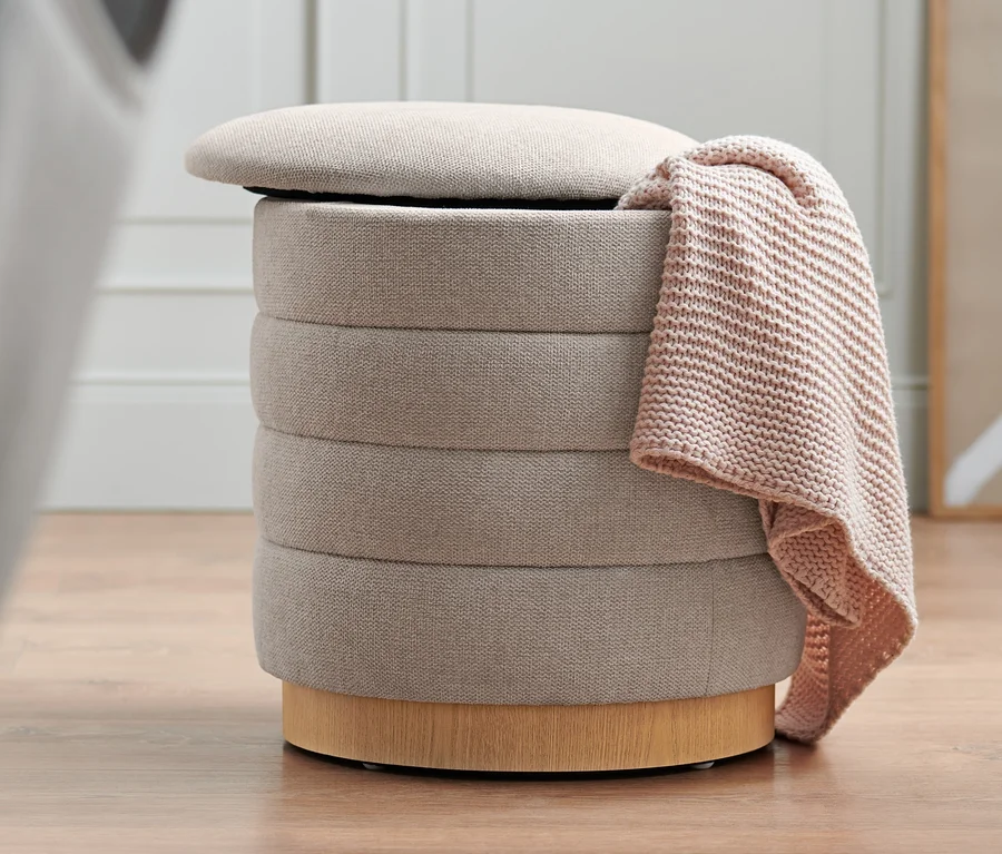Beiger 2-in-1-Pouf mit Stauraum, geöffnetem Deckel und einer Decke darüber.