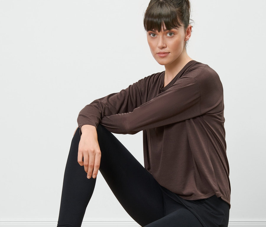 Frau sitzt auf einem Holzhocker und trägt ein braunes 2-in-1-Sportshirt und schwarze Leggings.