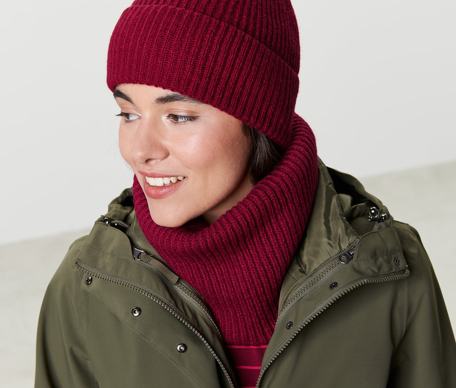 Frau schliesst olivgrüne Jacke und trägt einen roten Strickschlauchschal und eine Mütze.