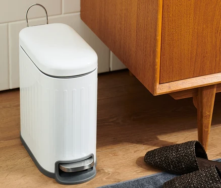 Im Badezimmer steht ein weißer Abfalleimer neben einem braunen Holzschrank und Hausschuhen auf einem grauen Teppich.
