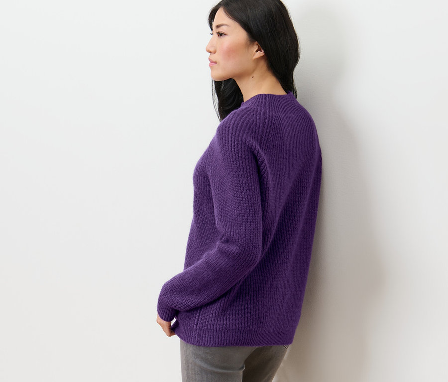 Seitenansicht einer Frau mit dunklen Haaren in einem lila Grobstrickpullover mit Stehkragen.