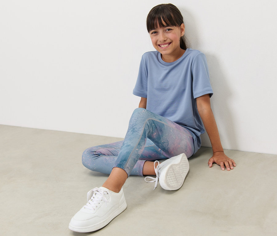 Mädchen sitzt am Boden und trägt eine Kinder-Sporttight mit Print und ein blaues T-Shirt.