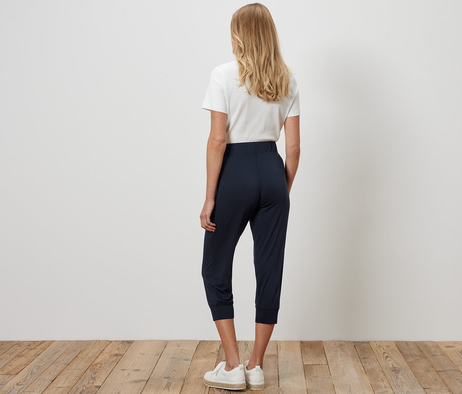 Frau von hinten stehend, trägt eine dunkelblaue 3/4-Jerseyhose, ein weißes T-Shirt und Turnschuhe.