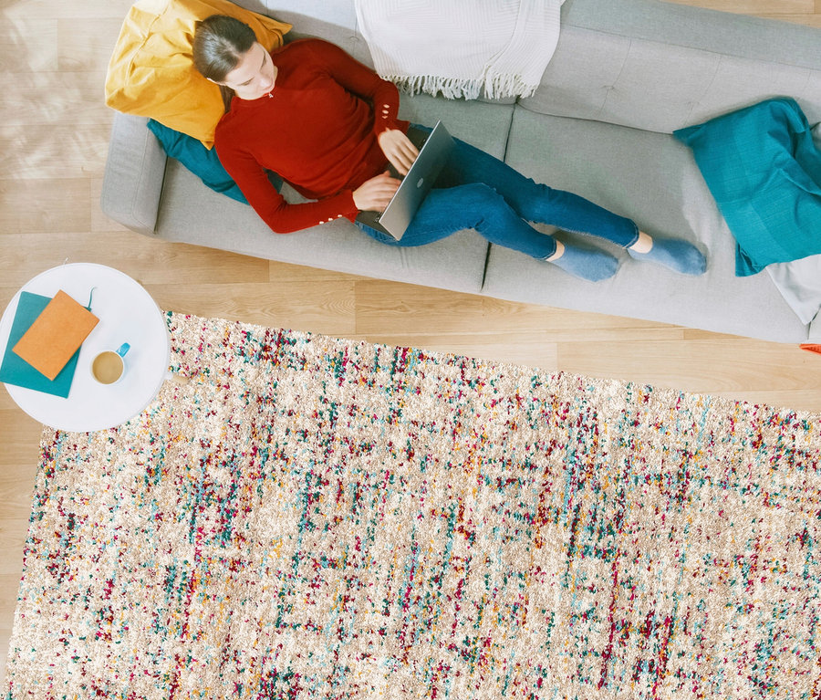Frau liegt auf grauem Sofa und benutzt Laptop. Darunter liegt ein beiger Teppich »Relic« mit gedämpften Pastellfarben.