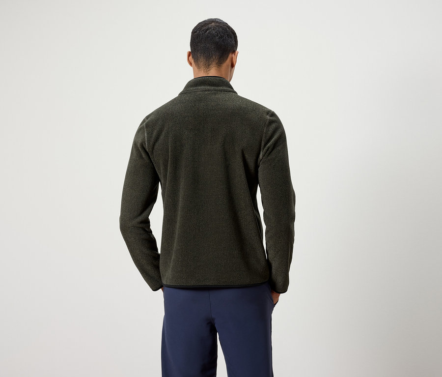 Ein Mann mit dem Rücken zur Kamera trägt eine olivgrüne Fleecejacke und dunkelblaue Hosen.