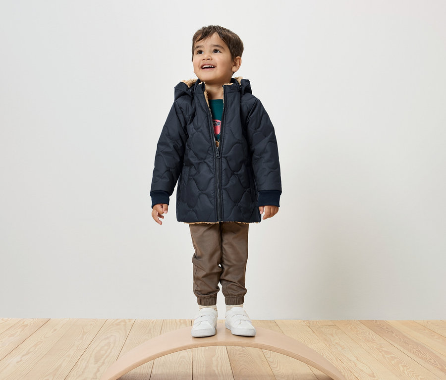 Ein Junge steht auf einem Balance Board und trägt eine marineblaue Steppjacke, braune Hosen und weiße Turnschuhe.