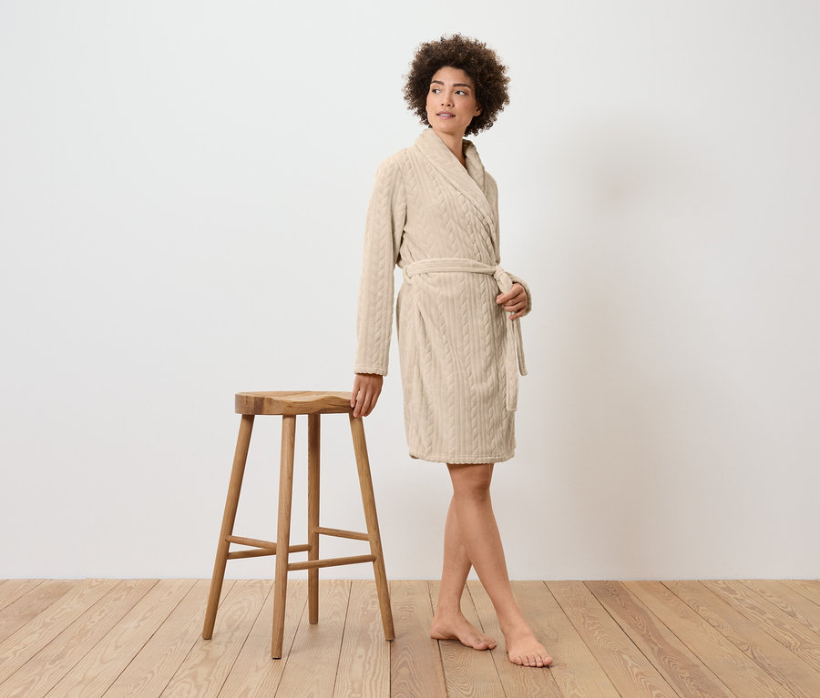 Frau im cremefarbenen Bademantel steht neben einem Holzhocker.