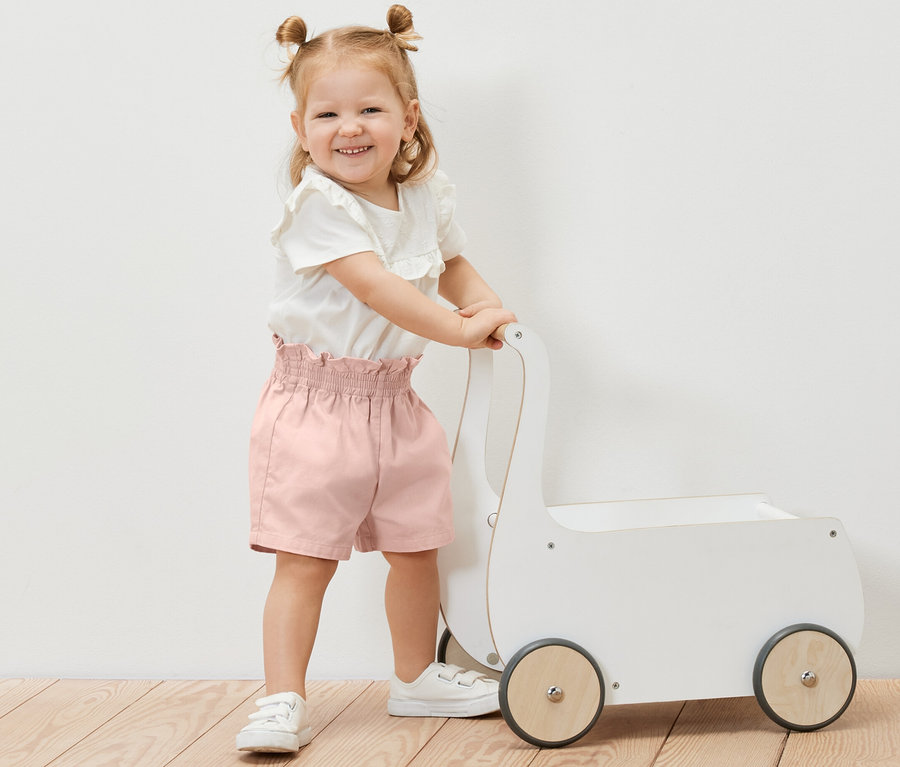 Lächelndes kleines Mädchen schiebt einen weißen Spielzeugwagen und trägt eine Kinder-Paperbag-Shorts.