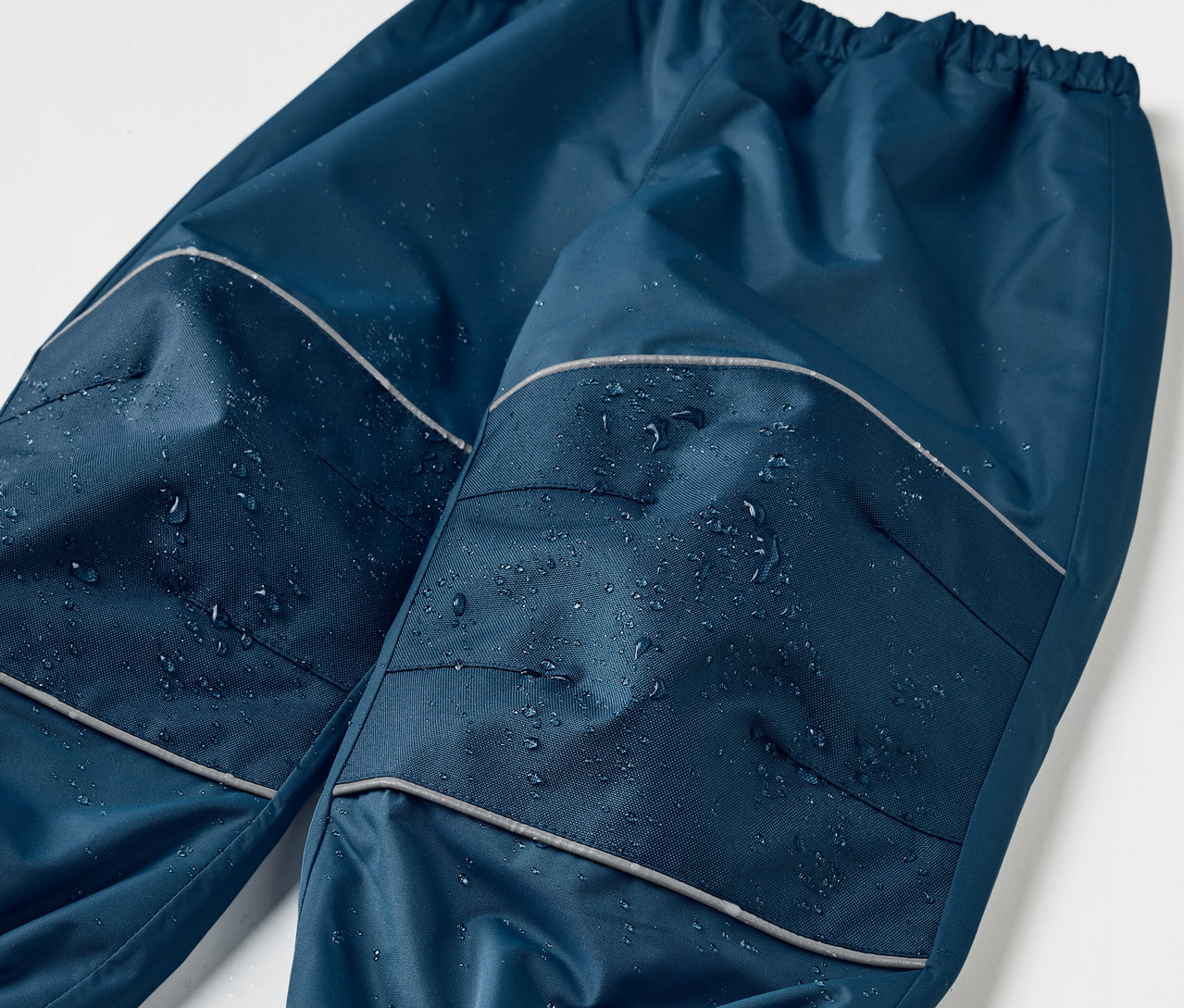 Detailaufnahme einer dunkelblauen Kinder-Regenhose mit Mitwachsfunktion, Reflexdetails und Wassertropfen.