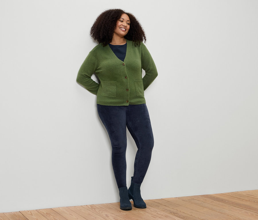 Frau mit lockigem Haar lehnt an einer Wand. Sie trägt eine grüne Strickjacke, blaue Leggings und Stiefel.