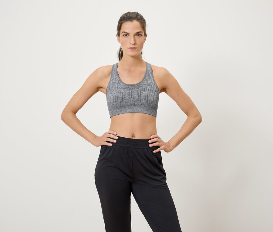 Frau posiert vor weißem Hintergrund in Sport-BH und schwarzer Hose.