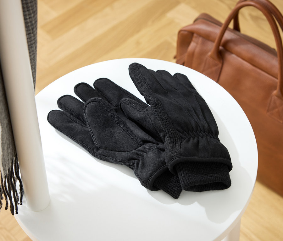 Auf einem weißen Hocker liegen schwarze Handschuhe, daneben eine braune Ledertasche und ein grauer Schal.