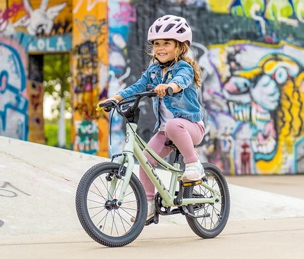 Ein junges Mädchen mit rosa Helm fährt ein mintgrünes PUKY LS-PRO 16 Fahrrad vor einer Wand mit Graffiti.