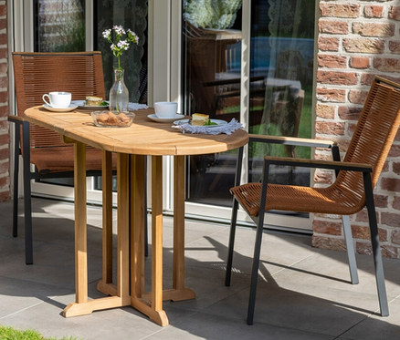 Zwei Korbstühle stehen an einem deVries Teak Balkontisch auf einer Terrasse mit Backsteinmauer.