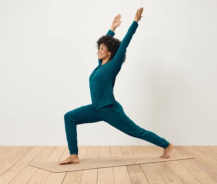 Frau mit lockigem Haar in blaugrüner Kleidung macht Yoga auf einer Matte.