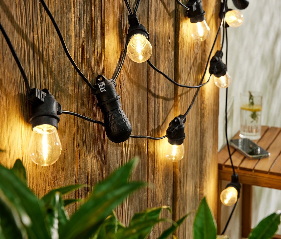 LED-Outdoor-Lichterkette mit Bluetooth-Lautsprechern hängt an einer Holzwand. Auf dem Tisch stehen ein Getränk und ein Handy.